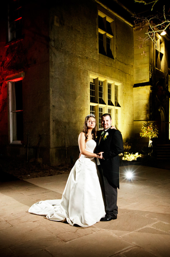 A wedding couple outside with Burton Court illuminated in the background in red and yellow.